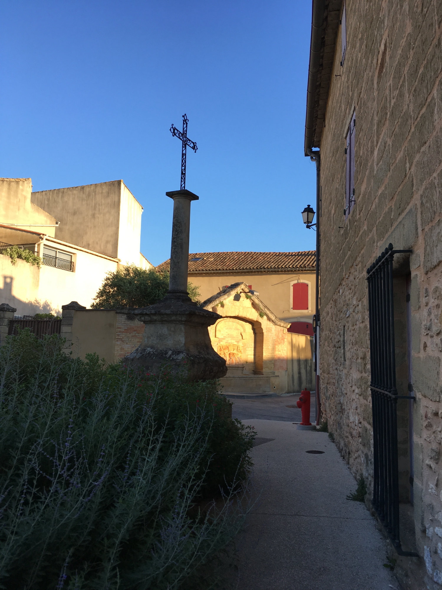 Narrow village lane in Uzès area with ornate iron cross on stone pillar, arched gateway, and warm-lit limestone buildings at dusk.